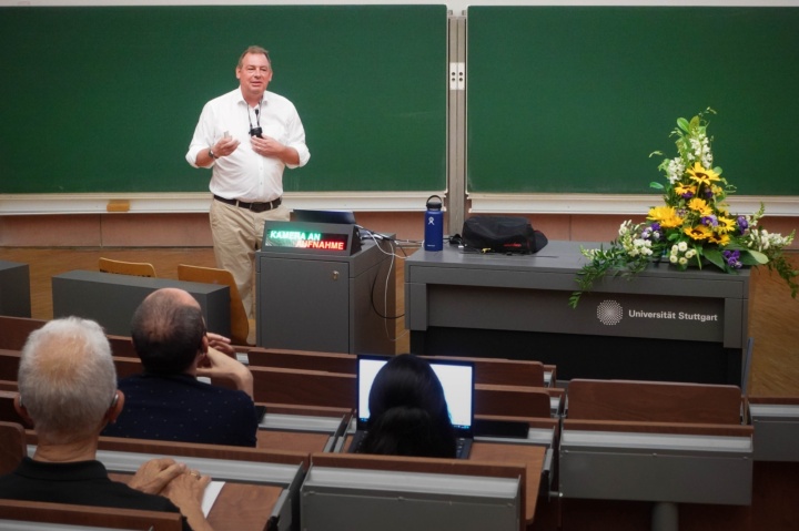 Prof. Becker im weißen Hemd spricht im Hörsaal vor einer grünen Tafel. Pult ist mit Blumengedeck geschmückt.