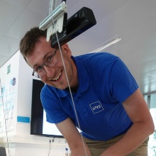 Mann mit blauem IPVS-Shirt steht gebeugt hinter Halterung mit Kabel und lacht in die Kamera.
